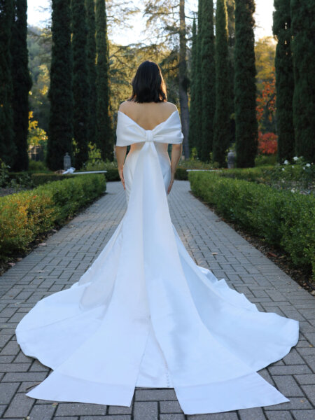 Essense of Australia D3754 off the shoulder mermaid wedding dress in plain fabric with stunning statement bow and train full length back view.
